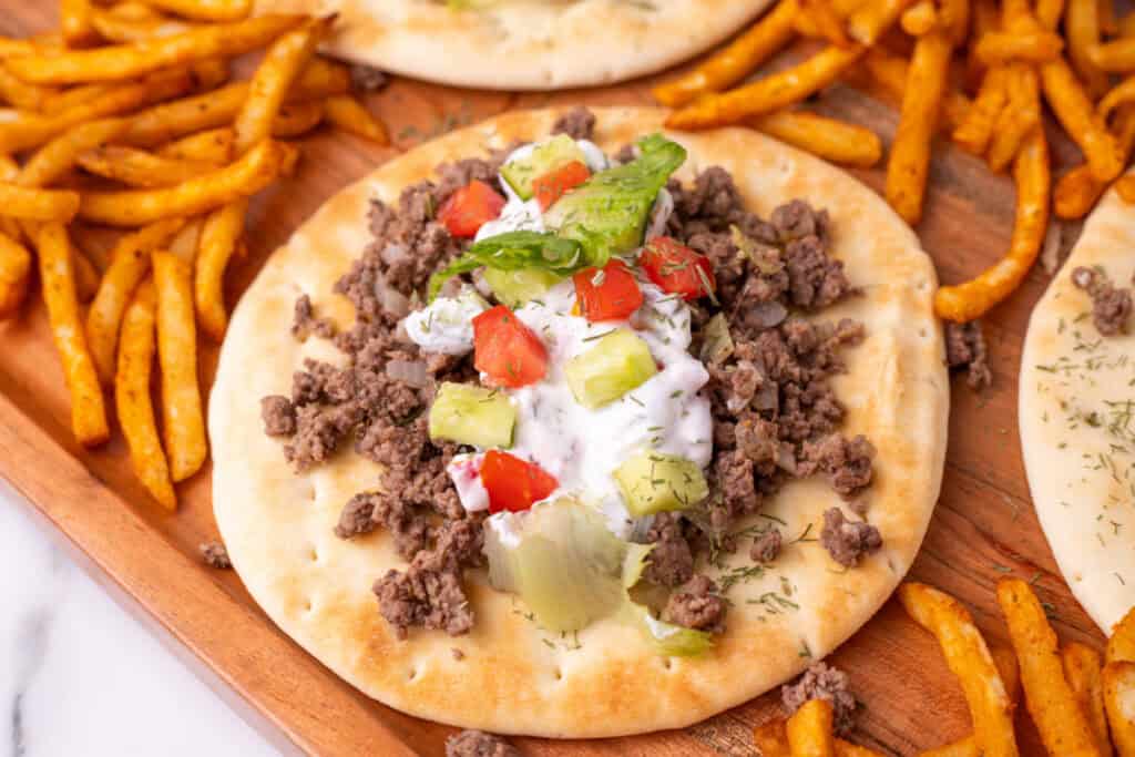 close view of pita bread topped with ground beef, yogurt sauce, cucumber, tomato, and lettuce on a wooden tray