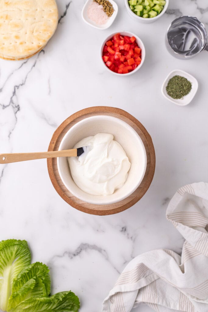 bowl of plain Greek yogurt with a spatula on a marble surface surrounded by chopped vegetables and pita bread