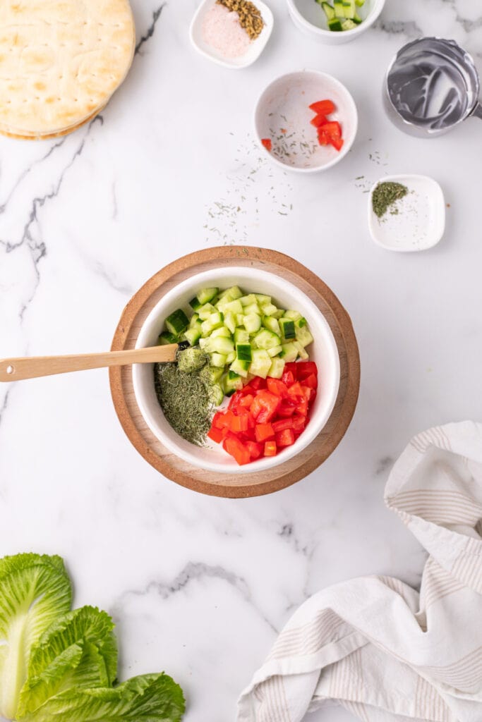 bowl with chopped cucumber, tomato, and dried dill being added to Greek yogurt