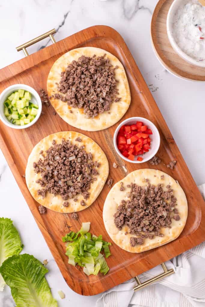 pita breads topped with ground beef and yogurt sauce on a serving tray with chopped vegetables