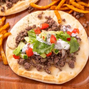pita breads topped with ground beef, yogurt sauce, cucumber, tomato, and lettuce on a wooden tray