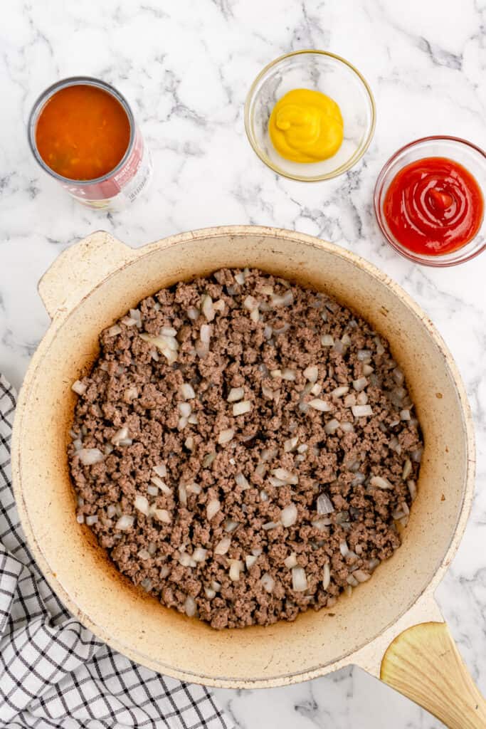 browned ground beef and diced onions cooking in a skillet with bowls of ketchup, mustard, and canned gumbo soup nearby