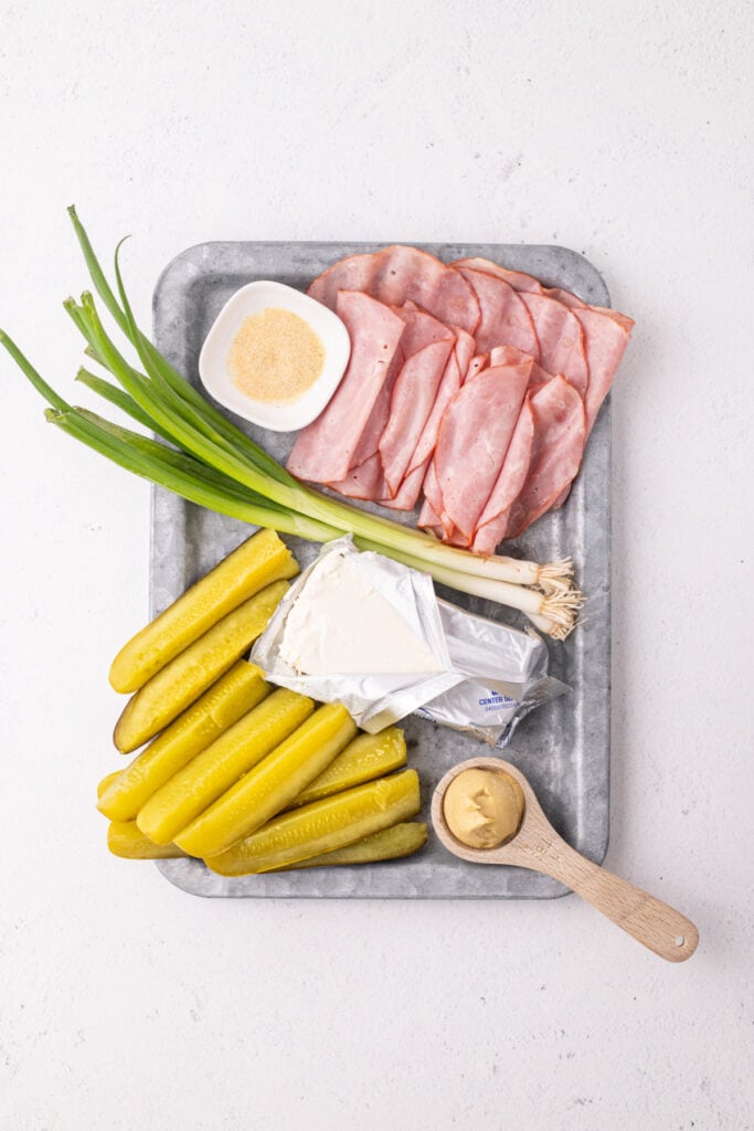 ingredients for Ham And Pickle Roll-Ups on a tray including sliced ham, dill pickles, cream cheese, mustard, garlic powder, and green onions