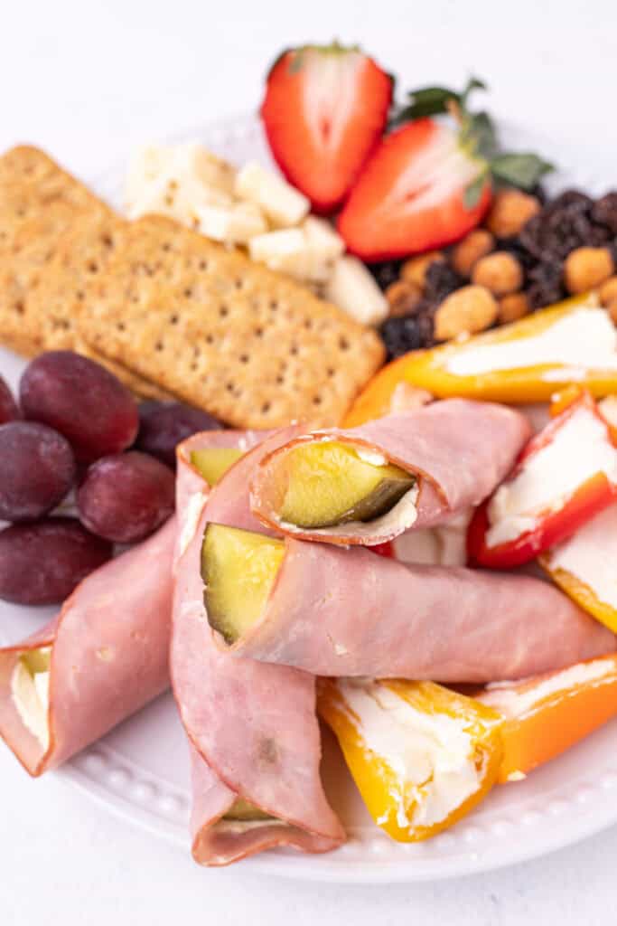 Ham And Pickle Roll-Ups served on a plate with crackers, grapes, strawberries, and cheese