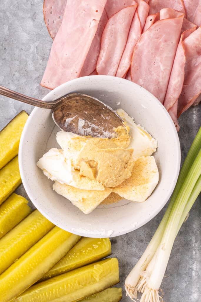 cream cheese, mustard, and garlic powder in a bowl with sliced ham and pickles nearby