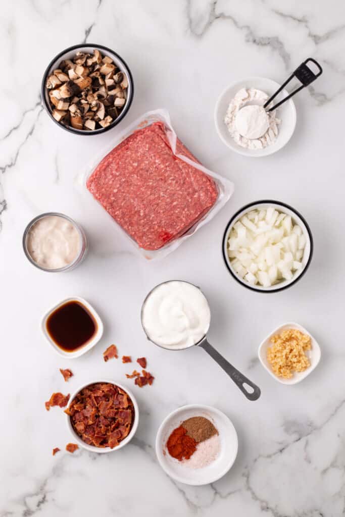 raw ground beef, chopped mushrooms, diced onion, sour cream, bacon, flour, garlic, Worcestershire sauce, and spices arranged on a white marble surface for hamburger stroganoff sloppy joes
