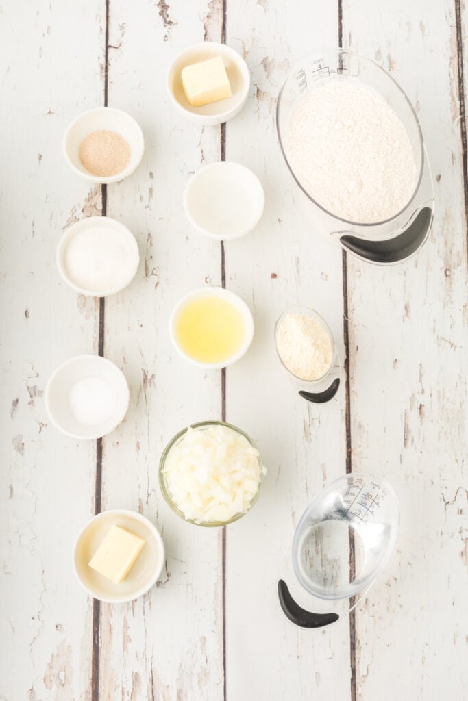 ingredients for homemade onion rolls including flour, butter, chopped onion, yeast, egg white, potato flakes, salt, sugar, and water on a white wooden surface