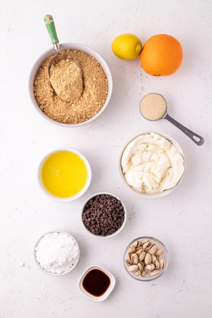 graham cracker crumbs, ricotta, chocolate chips, pistachios, powdered sugar, melted butter, vanilla, lemon, and orange on a white surface