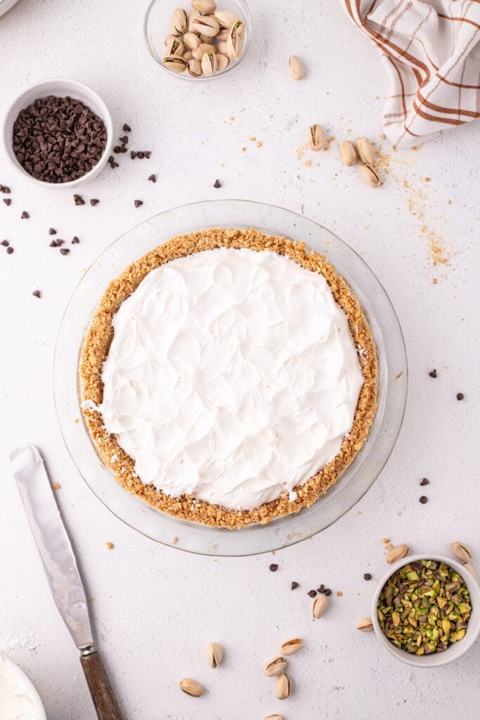 Overhead view of a no-bake cannoli pie with whipped cream fully spread across the top in a glass pie dish, surrounded by mini chocolate chips, pistachios, and a linen towel.
