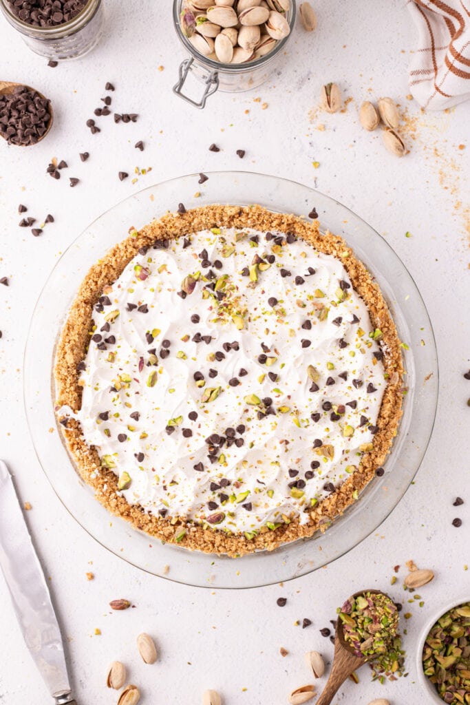 Overhead view of a finished no-bake cannoli pie topped with whipped cream, mini chocolate chips, and chopped pistachios in a glass pie dish, with a jar of pistachios and bowls of toppings around it.