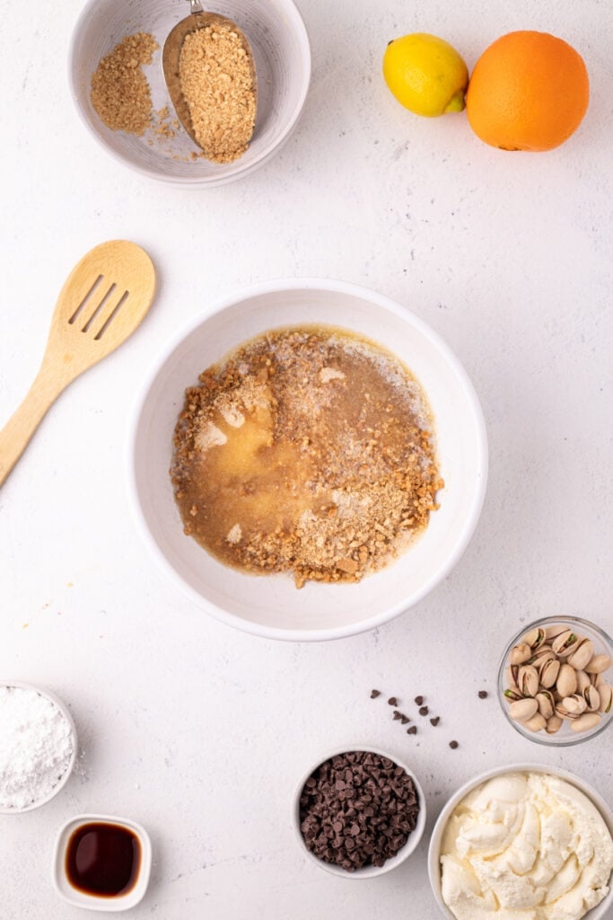 Overhead view of a white bowl with graham cracker crumbs and melted butter, surrounded by ingredients including a lemon, orange, mini chocolate chips, pistachios, powdered sugar, vanilla extract, and ricotta cheese on a white surface.