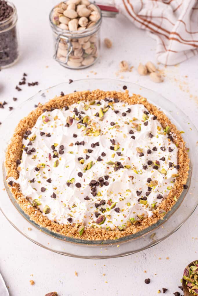 Angled view of a finished no-bake cannoli pie in a glass pie dish, topped with whipped cream, mini chocolate chips, and chopped pistachios, with a jar of pistachios and a striped linen towel in the background.