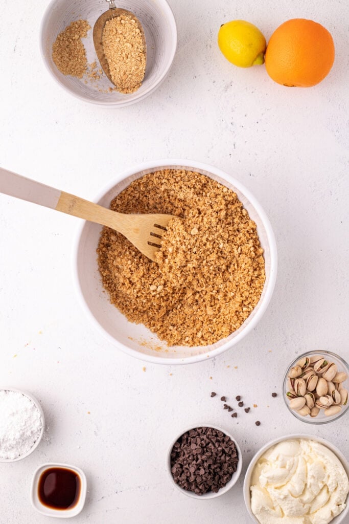 Overhead view of graham cracker crumbs mixed with butter in a white bowl with a wooden fork, surrounded by cannoli pie ingredients including a lemon, orange, chocolate chips, pistachios, powdered sugar, vanilla, and ricotta cheese.