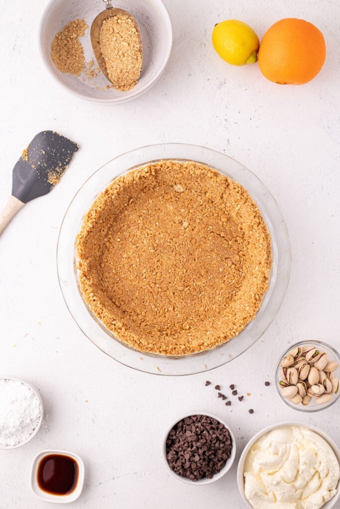 Overhead view of a pressed graham cracker crust in a glass pie dish, with a spatula nearby and surrounding ingredients including a lemon, orange, chocolate chips, pistachios, powdered sugar, vanilla extract, and ricotta cheese.