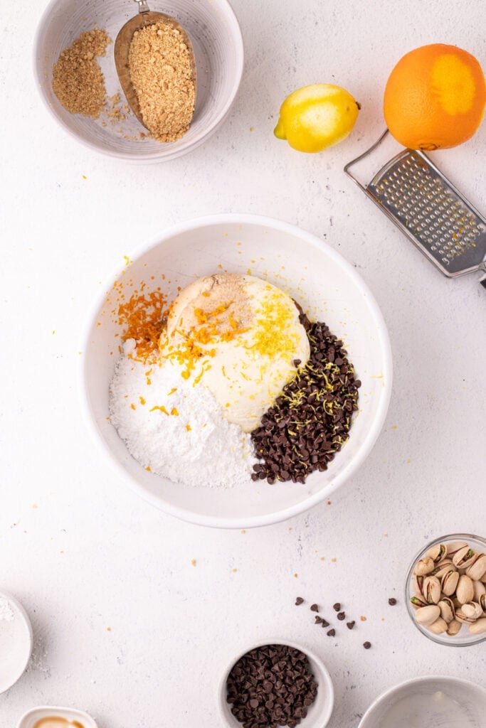 Overhead view of a white bowl with ricotta cheese, powdered sugar, mini chocolate chips, and fresh lemon and orange zest, with a box grater and whole citrus fruits nearby.