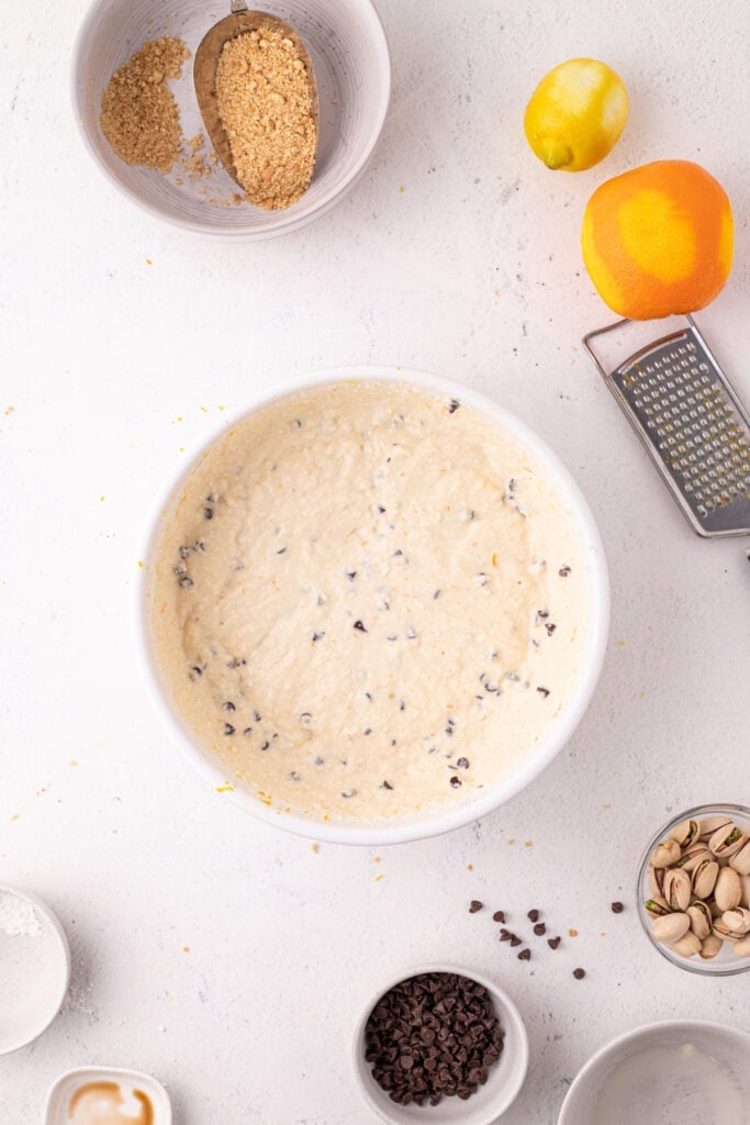 Overhead view of the smooth, creamy cannoli filling with mini chocolate chips mixed in, in a white bowl, with a lemon, orange, box grater, pistachios, and chocolate chips arranged around it.