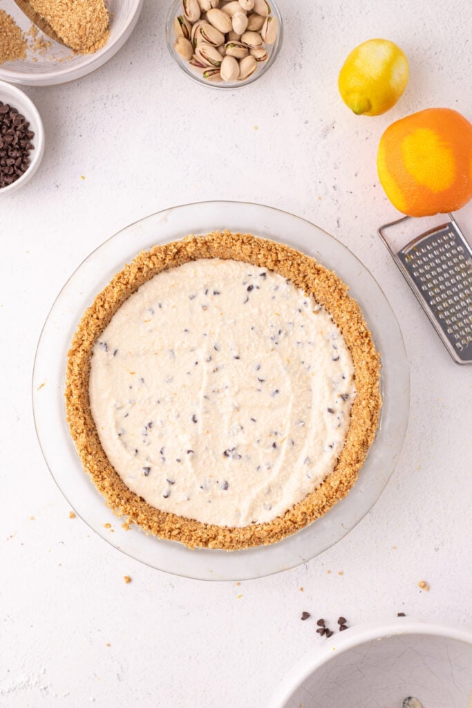 Overhead view of creamy cannoli filling spread into a graham cracker crust in a glass pie dish, with a lemon, orange, box grater, pistachios, and mini chocolate chips surrounding it.