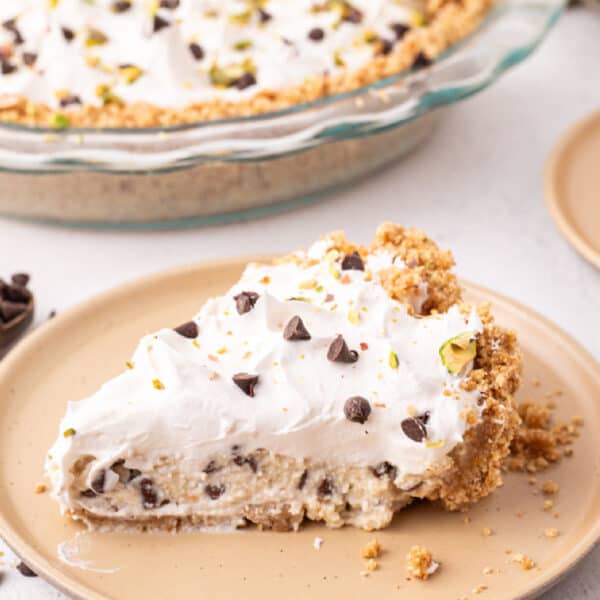 square cropped image of a slice of no bake cannoli pie on a plate with whipped topping, chocolate chips, and pistachios