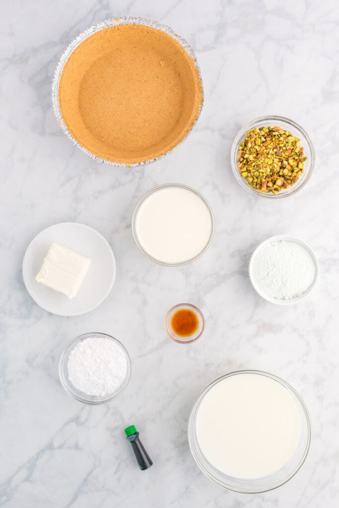 premade graham cracker crust, bowls of chopped pistachios, milk, powdered sugar, cream cheese, vanilla extract, and food coloring on a marble surface