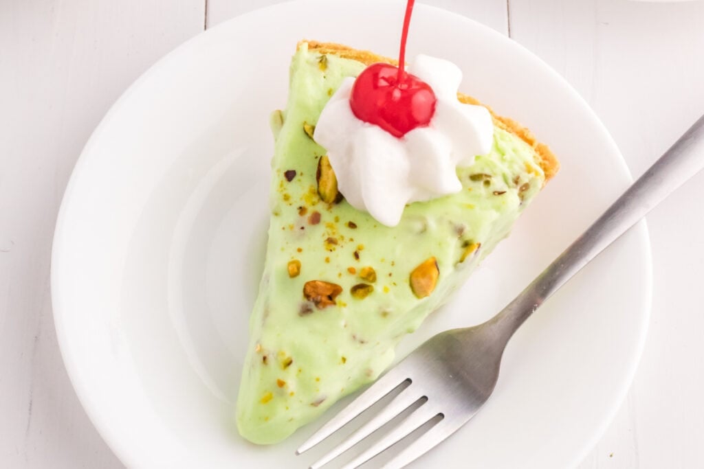 slice of pistachio cream pie topped with whipped cream and a cherry on a white plate