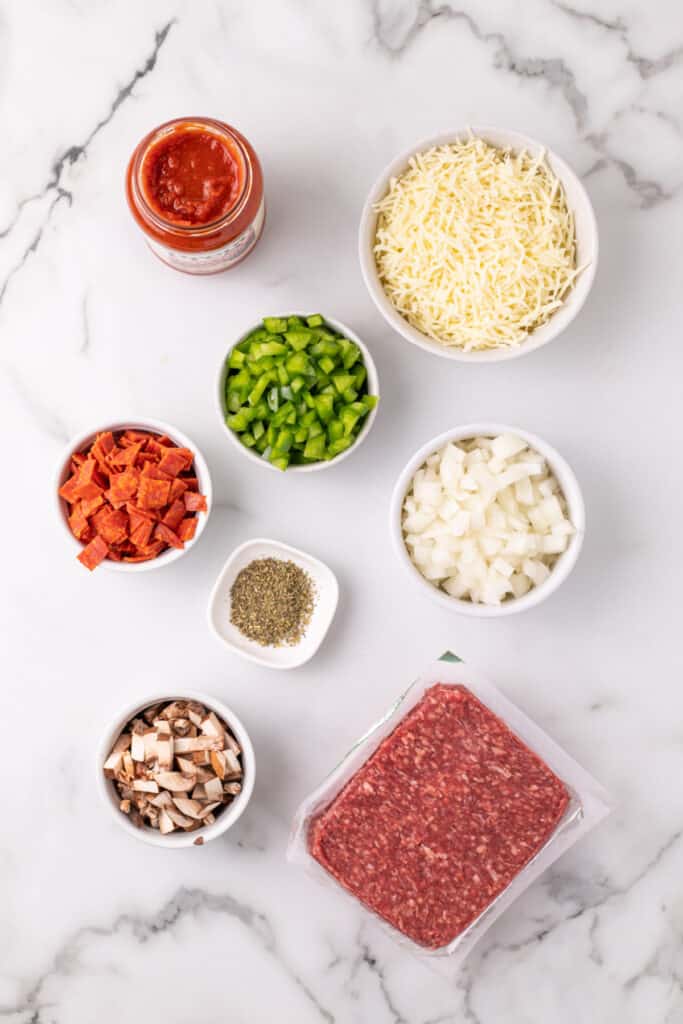 ingredients for Pizza Sloppy Joes including ground beef, chopped onion, green pepper, mushrooms, pepperoni, shredded mozzarella, pizza sauce, and dried oregano on a white surface