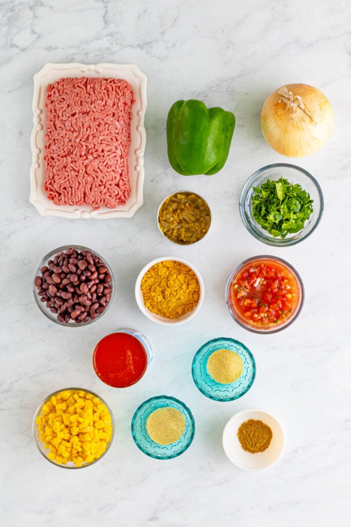Taco Sloppy Joes ingredients including ground beef, onion, green bell pepper, black beans, corn, salsa, tomato sauce, spices, and cilantro on a white surface