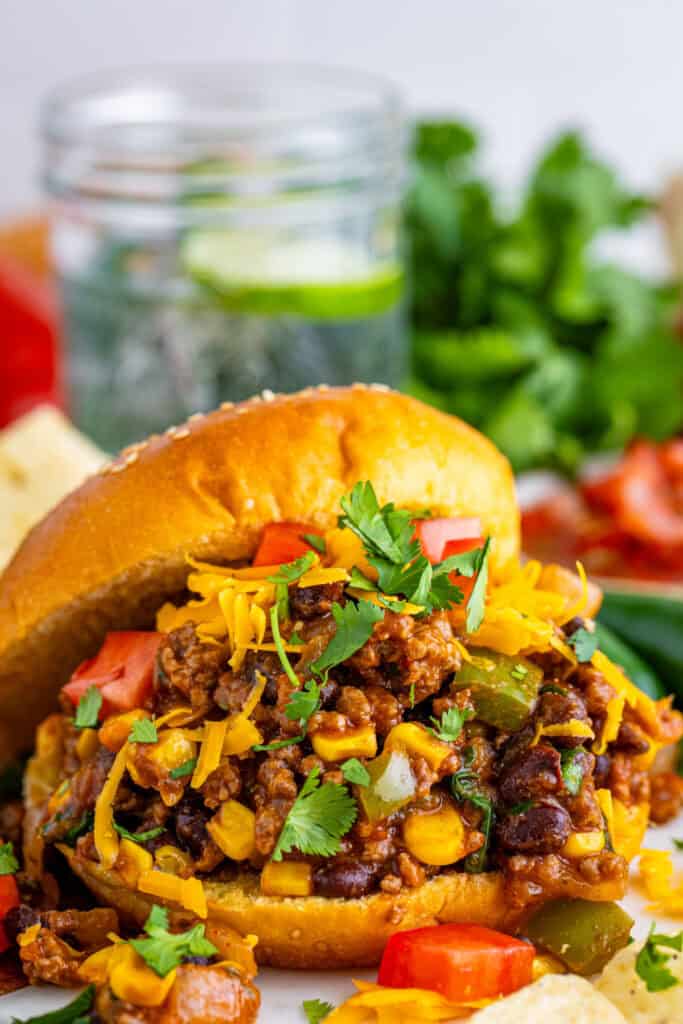 Taco Sloppy Joes sandwich on a bun topped with shredded cheese, tomatoes, and cilantro with chips on the side