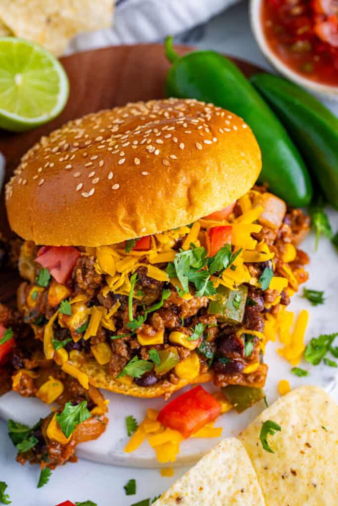 Taco Sloppy Joes sandwich with sesame seed bun, ground beef, black beans, corn, cheese, and cilantro served with chips and jalapenos