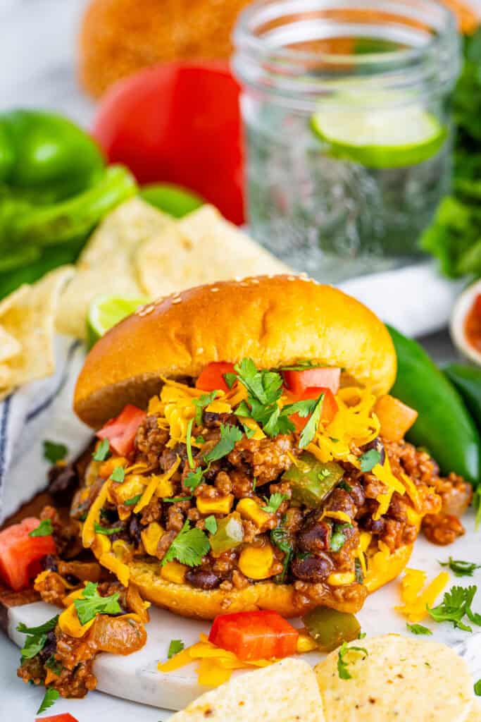 Taco Sloppy Joes sandwich on a bun topped with cheese, tomatoes, and cilantro served with chips