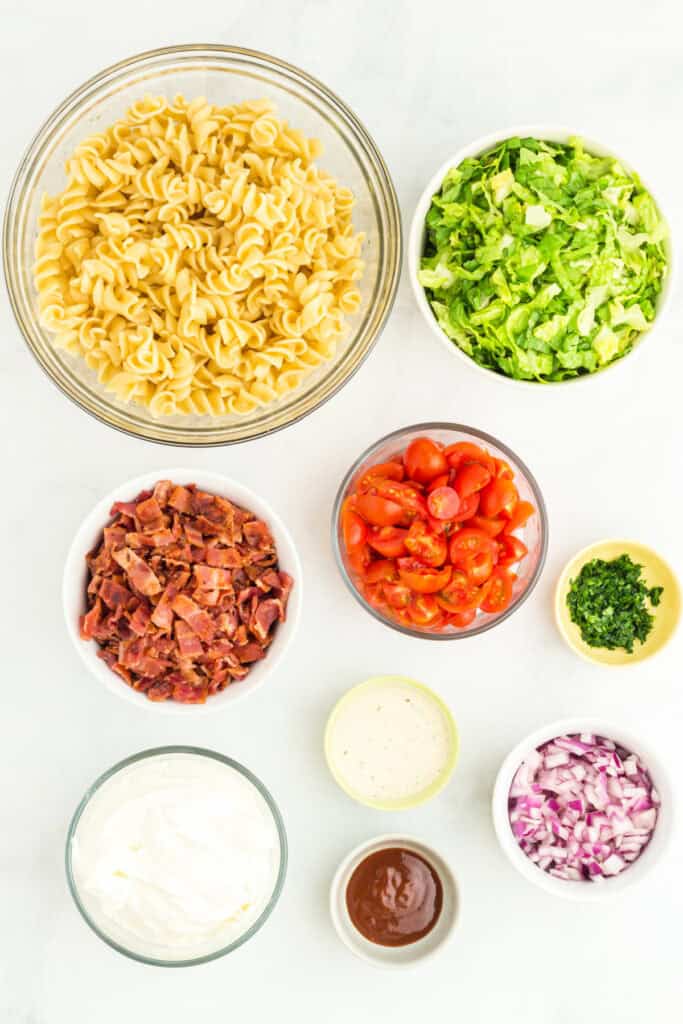 ingredients for BBQ Ranch BLT Pasta Salad including cooked rotini pasta, chopped bacon, shredded romaine lettuce, cherry tomatoes, red onion, sour cream, ranch seasoning, BBQ sauce, and parsley in bowls