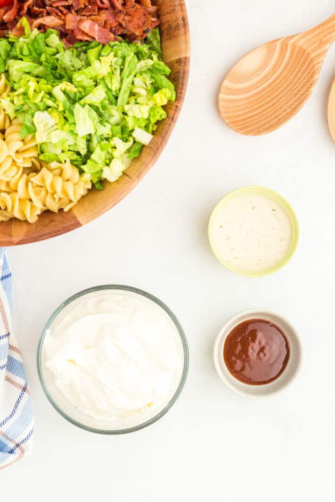 rotini pasta, shredded romaine lettuce, and chopped bacon in a wooden bowl with sour cream, ranch seasoning, and BBQ sauce nearby for BBQ Ranch BLT Pasta Salad