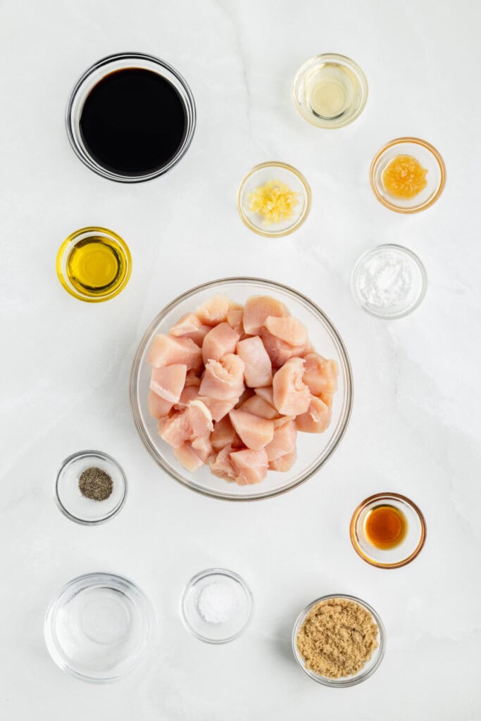 Ingredients for Chicken Teriyaki arranged on a white surface with raw chicken, soy sauce, garlic, ginger, brown sugar, and sesame oil