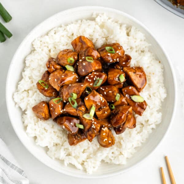 Chicken Teriyaki served over white rice with sesame seeds and sliced green onions in a white bowl