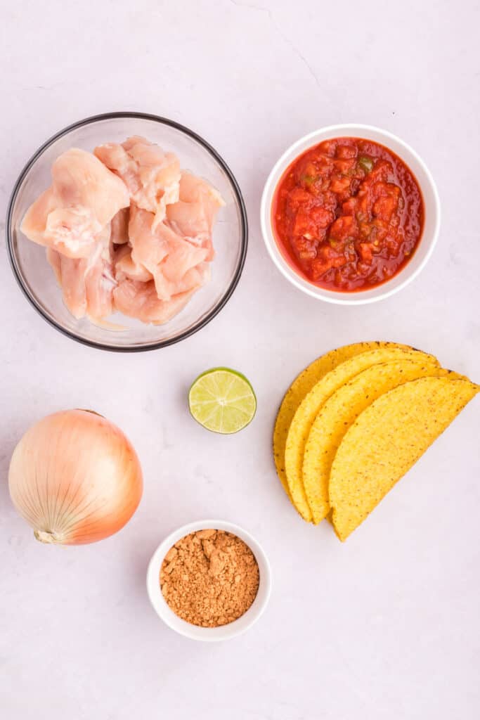 Ingredients for Crockpot Shredded Chicken Tacos including raw chicken, salsa, taco seasoning, onion, lime, and taco shells on a light background
