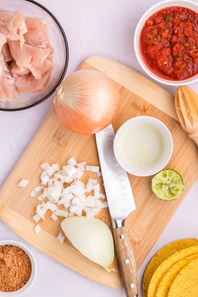 Chopped onion, lime juice, salsa, and raw chicken prepared for Crockpot Shredded Chicken Tacos on a cutting board