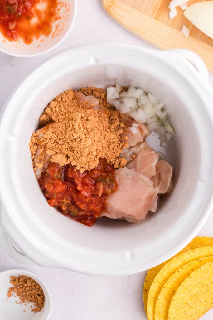 Raw chicken, salsa, onion, and taco seasoning added to a slow cooker for Crockpot Shredded Chicken Tacos