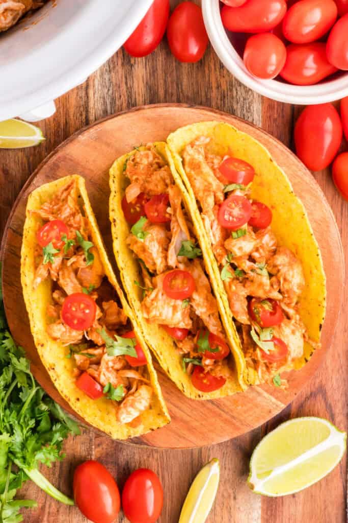 Three Crockpot Shredded Chicken Tacos on a wooden plate with cherry tomatoes, cilantro, and lime wedges