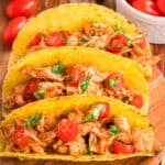 Square image of Crockpot Shredded Chicken Tacos topped with cherry tomatoes and cilantro in crispy taco shells
