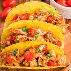 Square image of Crockpot Shredded Chicken Tacos topped with cherry tomatoes and cilantro in crispy taco shells