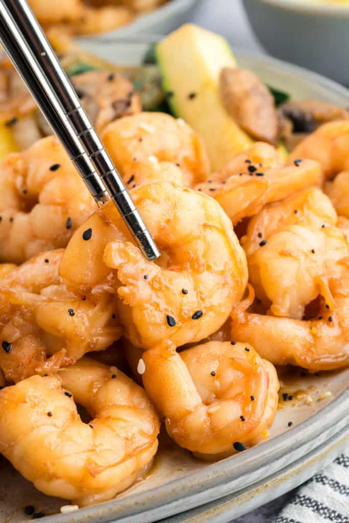 Chopsticks holding a piece of Hibachi Shrimp above a plate of shrimp and hibachi vegetables.