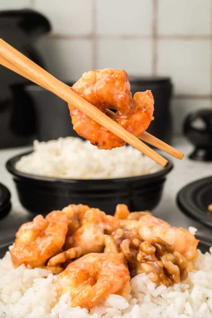 Chopsticks holding a piece of Honey Walnut Shrimp above white rice and candied walnuts
