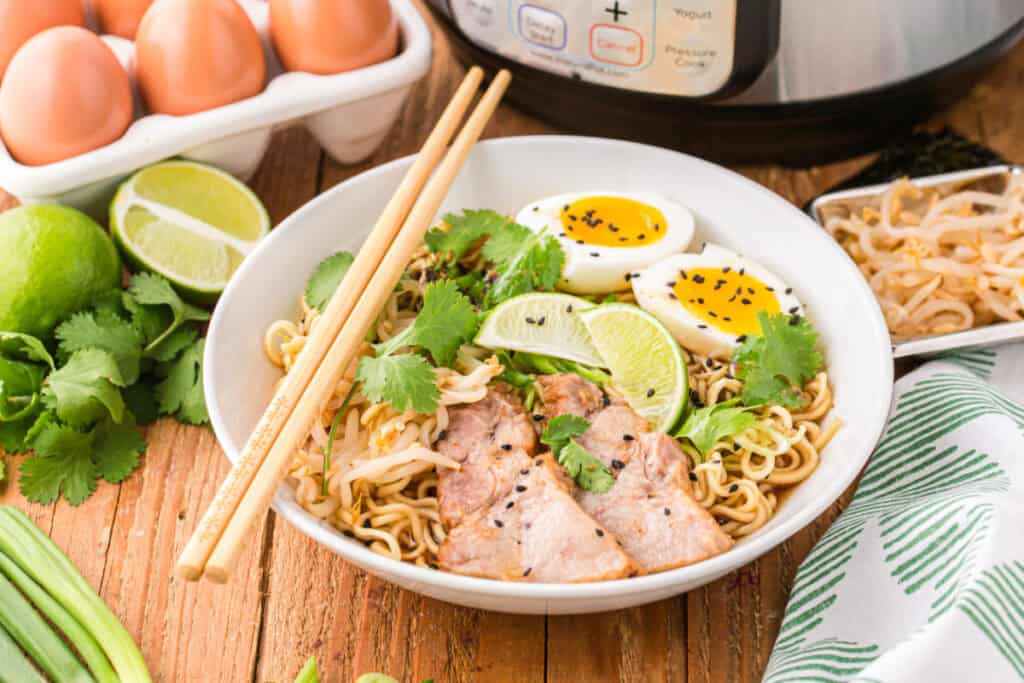 Bowl of Instant Pot Ramen topped with sliced pork, soft-boiled eggs, bean sprouts, cilantro, lime wedges, and black sesame seeds with chopsticks resting across the bowl
