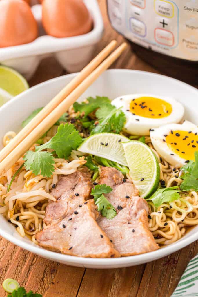 Close-up of Instant Pot Ramen with sliced pork, soft-boiled eggs, bean sprouts, cilantro, lime wedges, black sesame seeds, and chopsticks