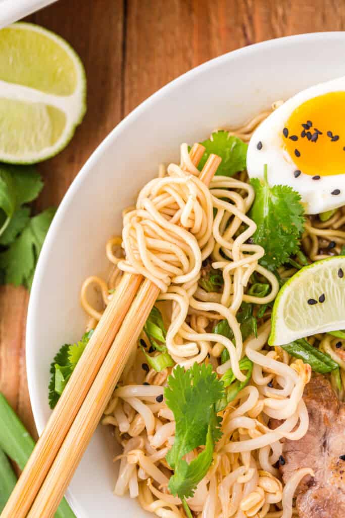 Close-up of chopsticks lifting ramen noodles from a bowl of Instant Pot Ramen topped with soft-boiled egg, bean sprouts, cilantro, green onions, lime wedge, and sliced pork