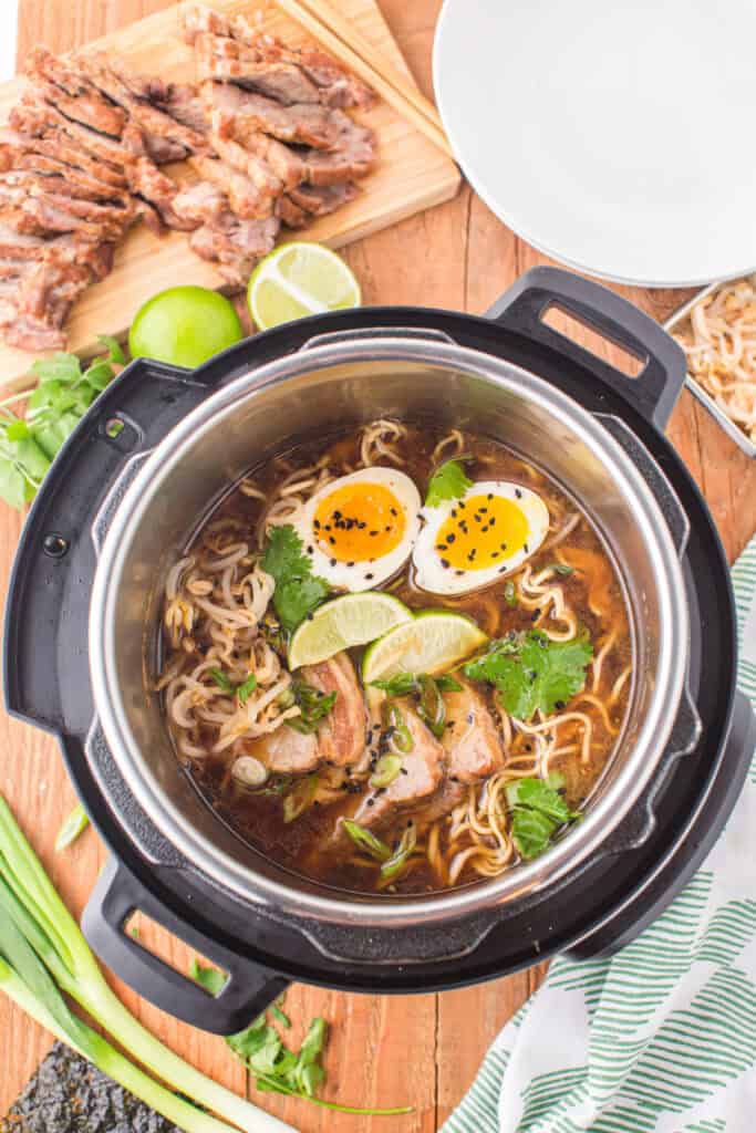 Instant Pot filled with ramen broth, noodles, sliced pork, soft-boiled eggs, cilantro, green onions, bean sprouts, lime wedges, and black sesame seeds