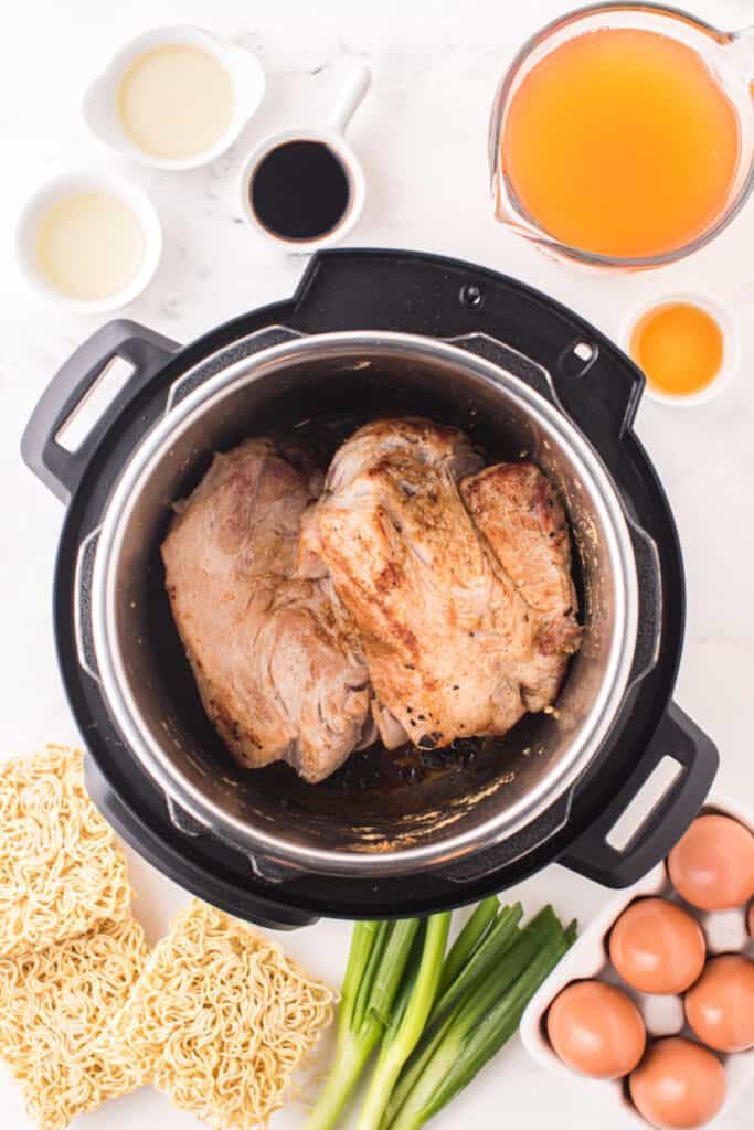 Browned pork shoulder in an Instant Pot surrounded by ramen noodles, eggs, green onions, and broth ingredients