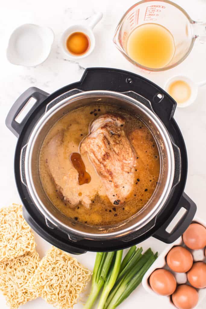 Browned pork shoulder in broth inside an Instant Pot with ramen noodles, eggs, and green onions nearby