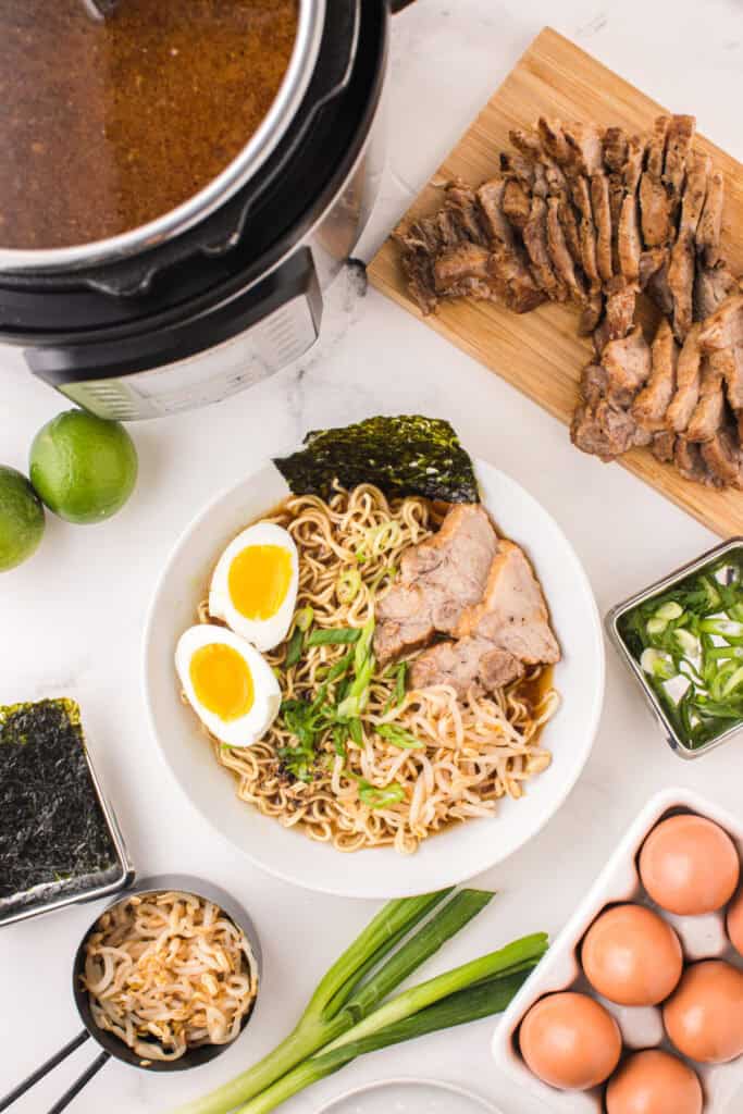Bowl of Instant Pot Ramen topped with sliced pork, soft-boiled eggs, bean sprouts, green onions, and nori beside an Instant Pot