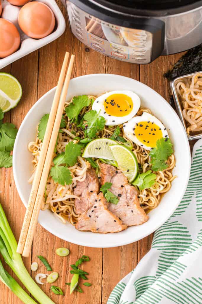 Bowl of Instant Pot Ramen topped with sliced pork, soft-boiled eggs, bean sprouts, cilantro, green onions, lime wedges, and black sesame seeds