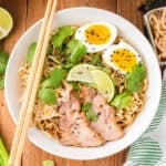 Square image of Instant Pot Ramen topped with sliced pork, soft-boiled eggs, bean sprouts, cilantro, lime wedges, black sesame seeds, and chopsticks
